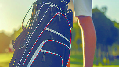 Ein Golfer steht neben einer schwarzen Golfbag mit mehreren Schlägern auf dem Rasen.