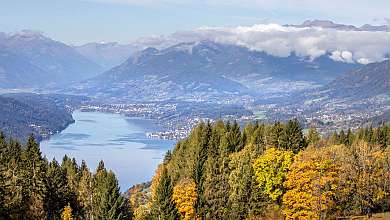 Blick über einen Bergsee mit Wäldern, herbstlichen Bäumen und nebelverhangenen Bergen.