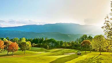 Weitläufiger Golfplatz mit Bäumen, Hügeln, Golfwagen und Bergpanorama im Abendlicht.
