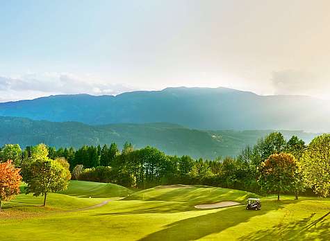 Weitläufiger Golfplatz mit Hügeln, Bäumen und Bergen im Hintergrund.