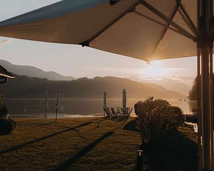 Terrasse am See mit Sonnenschirmen, Stühlen und Blick auf Berge im Abendlicht.