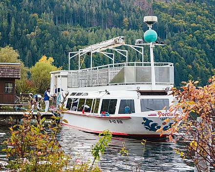 Weißes Ausflugsboot liegt an einem Steg vor bewaldeten Bergen.
