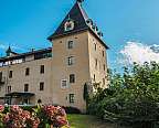 Historisches Gebäude mit Turm und Spitzdach, umgeben von Garten und blauem Himmel.
