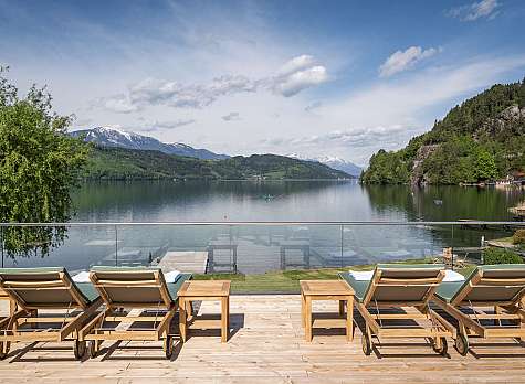 Holzterrasse mit Liegestühlen am See vor Bergen und blauem Himmel