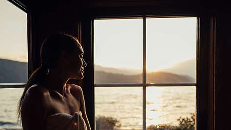 A person sits wrapped in a towel in front of a window overlooking the sea at sunset.