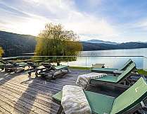 Holzterrasse mit grünen Liegen am ruhigen See vor Bergen und Himmel.