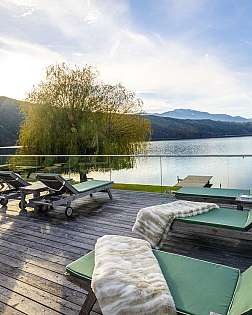 Holzterrasse mit grünen Liegen am ruhigen See vor Bergen und Himmel.