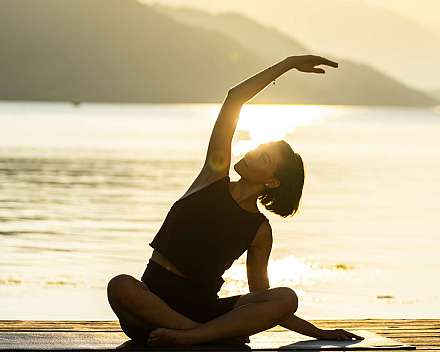 Eine Frau sitzt auf einer Matte am Steg und macht eine Yoga-Dehnung im Gegenlicht.