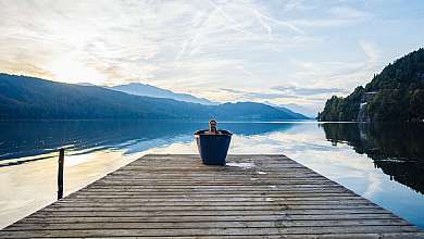 Eine Person sitzt in einem blauen Bottich auf einem Steg am ruhigen See.