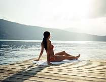 Woman in a bikini sitting on a wooden platform by the lake and looking out over the water.