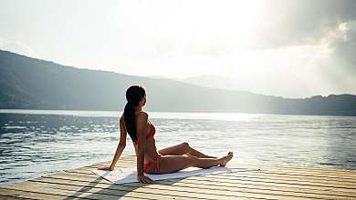 Woman in a bikini sitting on a wooden platform by the lake and looking out over the water.