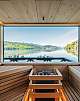 Wooden sauna with panoramic windows and a view of a calm lake and green mountains.