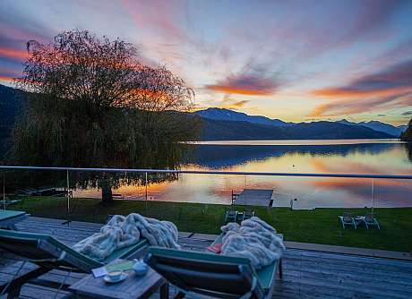 View from a terrace over a calm lake at sunset with mountains and sun loungers.