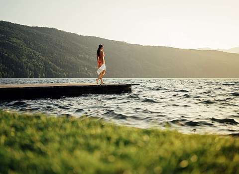 A woman stands on a wooden jetty by the lake in front of wooded mountains.