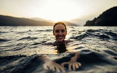 Eine lächelnde Frau schwimmt bei Sonnenuntergang im Wasser vor Bergsilhouette.