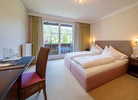 Helles Hotelzimmer mit Doppelbett, Schreibtisch und Balkonblick auf bewaldete Hügel.