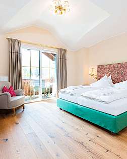 Helles Schlafzimmer mit großem Bett, Sessel, Holzkommode und Balkonfenster.