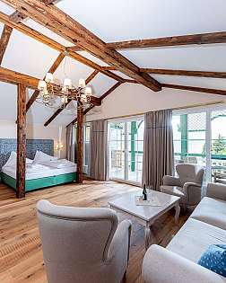 Geräumiges Hotelzimmer mit sichtbaren Holzbalken, Bett, Sitzbereich und großer Fensterfront.