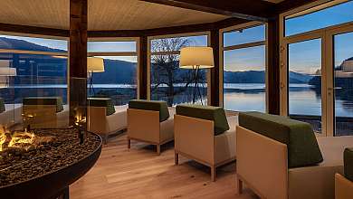 Gemütliche Lounge mit Sesseln, Kamin und großen Fenstern zum See und Bergen.
